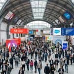 Thousands of digital marketing professionals filling the exhibition floor at DMEXCO 2026 in the Koelnmesse, Cologne, Germany, with branded booths and digital screens visible throughout the hall